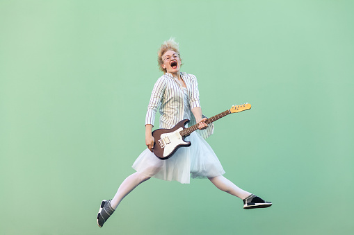 Portrait of young happy blonde woman in white shirt, skirt, and striped blouse with eyeglasses holding electric guitar, screaming and jumping. indoor studio shot isolated on light green background.
