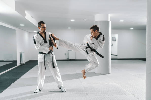 istock Young Caucasian boy in dobok kicking barefoot while trainer holding kick target. Taekwondo training concept. 1142167490