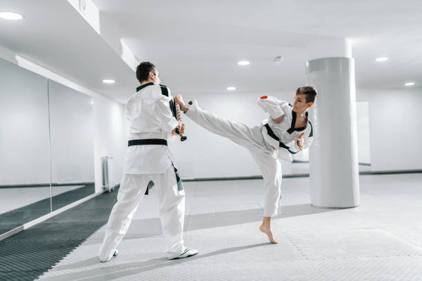 istock Young Caucasian boy in dobok kicking barefoot while trainer holding kick target. Taekwondo training concept. 1142167553