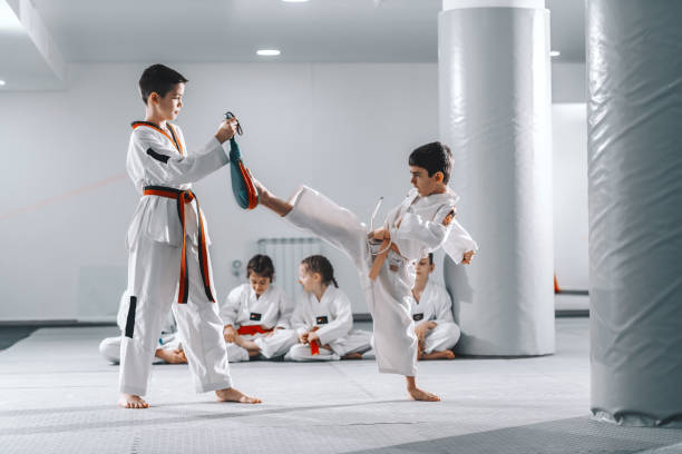 istock Two young Caucasian boys in doboks having taekwondo training at gym. One boy kicking while other one holding kick target. In background their friend sitting with legs crossed and watching them. 1142168917