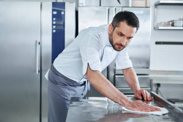 When preparing foods keep it clean, a dirty area should not be seen. Young male professional cook cleaning in commercial kitchen Bearded man prepares the surface for cooking in the kitchen. Cook carefully wipes the surface. Health and safety concept. commercial kitchen photos stock pictures, royalty-free photos & images