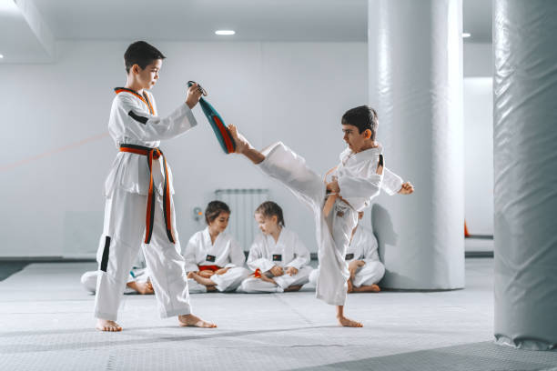 istock Two young Caucasian boys in doboks having taekwondo training at gym. One boy kicking while other one holding kick target. In background their friend sitting with legs crossed and watching them. 1142170241