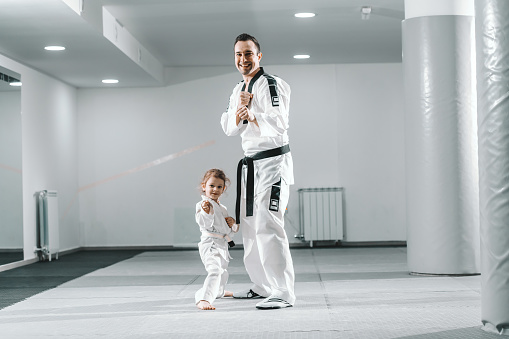 Smiling Caucasian taekwondo trainer posing with little girl in white gym.