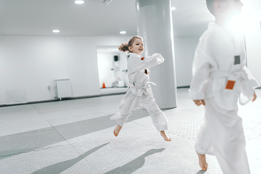 Smiling happy Caucasian little girl jumping at taekwondo training and wearing dobok.