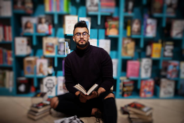 Smart arab student man in library Tall smart arab student man, wear on violet turtleneck and eyeglasses, at library sitting against books shelves. middle eastern ethnicity mature adult book reading stock pictures, royalty-free photos & images