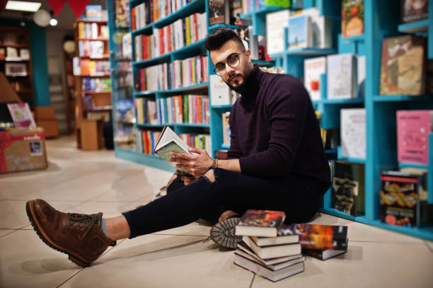 Smart arab student man in library Tall smart arab student man, wear on violet turtleneck and eyeglasses, at library sitting against books shelves. middle eastern ethnicity mature adult book reading stock pictures, royalty-free photos & images