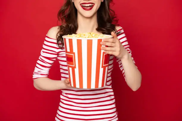 Facial expression concept. Waist up cropped head portrait of happy excited brunette woman staying with big box of pop corn isolated on red