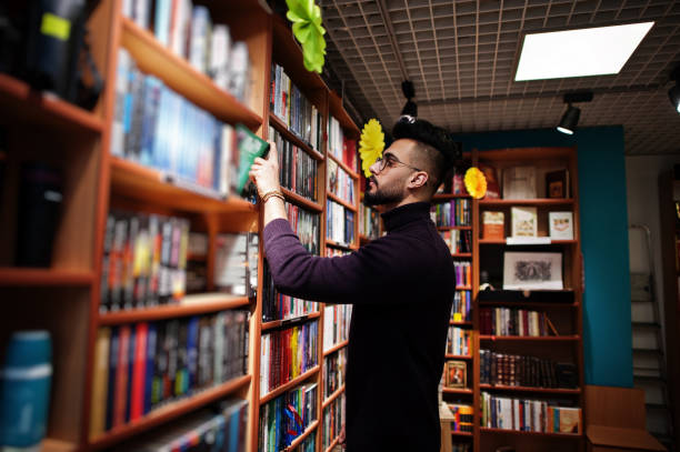 Smart arab student man in library Tall smart arab student man, wear on violet turtleneck and eyeglasses, at library selecting book on shelves. middle eastern ethnicity mature adult book reading stock pictures, royalty-free photos & images