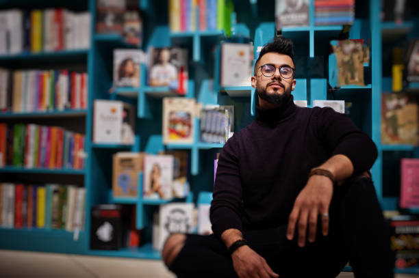 Smart arab student man in library Tall smart arab student man, wear on violet turtleneck and eyeglasses, at library sitting against book shelves. middle eastern ethnicity mature adult book reading stock pictures, royalty-free photos & images