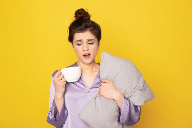 Sleepy girl with coffee and pillow yawning It's not easy to wake up. Waist up portrait of sleepy brunette female in silk pajamas with cup of coffee and pillow yawning. Isolated on yellow hair on pillow stock pictures, royalty-free photos & images