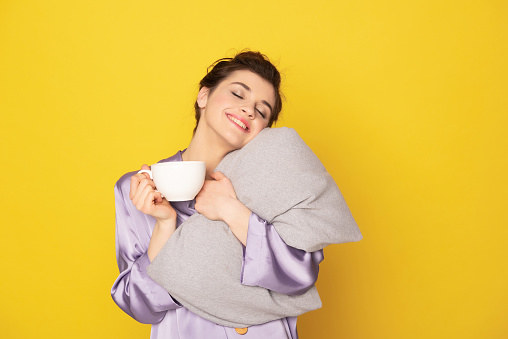 It's not easy to wake up. Waist up portrait of dreamful sleepy brunette female in silk pajamas staying with coffee and embracing pillow. Isolated on yellow