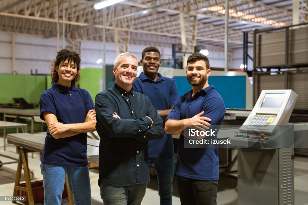 Confident workers standing in industry Portrait of smiling multi-ethnic workers in industry. Confident male instructor is with trainees. They are standing in factory. Manufacturing Occupation Stock Photo