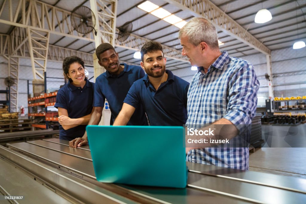 Instructor explaining trainees over laptop Male instructor explaining trainees over laptop. Confident apprentice are listening to engineer. They are standing in factory. Factory Stock Photo