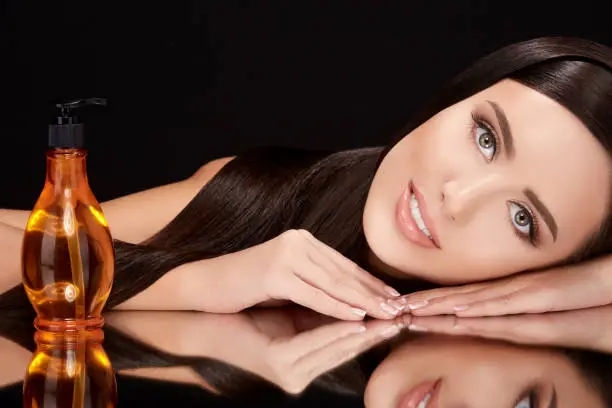 Beauty concept, brunette woman with light eyes lying on black glass with her reflexion. Gorgeous model with hair oil near her, head and shoulders, beauty portrait, closeup