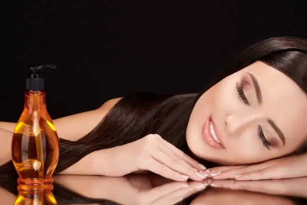 Beauty concept, brunette woman with closed eyes lying on black glass with her reflexion. Gorgeous model with hair oil near her, head and shoulders, beauty portrait, closeup