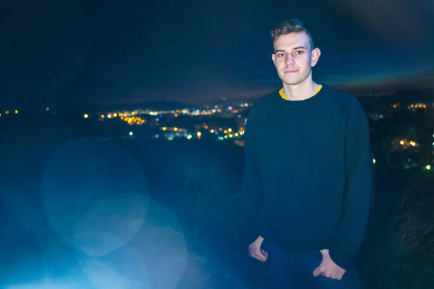 Young adult man by city views at dusk. He is wearing casual clothes and looking to the camera. Blue lighting effect into the camera.