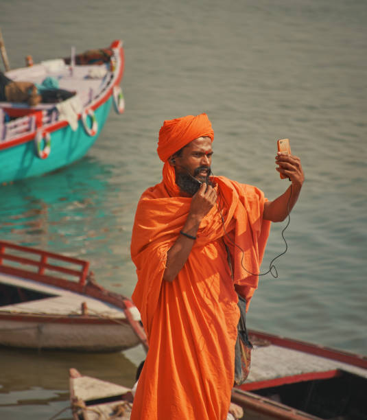 A sadhu, dressed in their traditional saffron coloured clothing, talking in his mobile, probably making a video call. He is standing at the ghats, boats floating in Ganges are seen behind.\nVaranasi@ Uttar Pradesh is one the oldest living cities of world and called spiritual capital of India. The city is on the banks of river Ganga. \nPhoto taken at Varanasi, Uttar Pradesh on 11/19/2018.