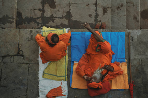 High angle shot of Two sadhus at Varanasi riverbanks, at Dashashwamedh ghat. One of them is sitting while other sadhu is taking a nap. Both are dressed in their traditional saffron clothes. Shot taken from a balcony above their head.\nVaranasi@ Uttar Pradesh is one the oldest living cities of world and called spiritual capital of India. The city is on the banks of river Ganga. \nPhoto taken at Varanasi, Uttar Pradesh on 11/19/2018.