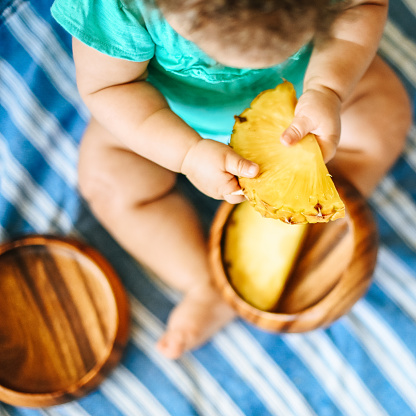 Little girl, 8 month old baby, holds cute up piece of pineapple in her cute little fingers, wooden bowls full of fresh fruit snack and a curious small child. Baby is mixed race half African American half Caucasian and she sits on a striped picnic blanket with the fresh ripe raw fruit