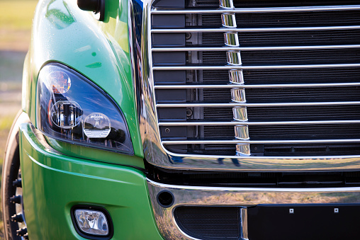 Sunlit stylish and comfortable green big rig semi truck of latest model of commercial long-distance transport with shiny chrome grille and efficient headlight in the parking lot waiting for cargo
