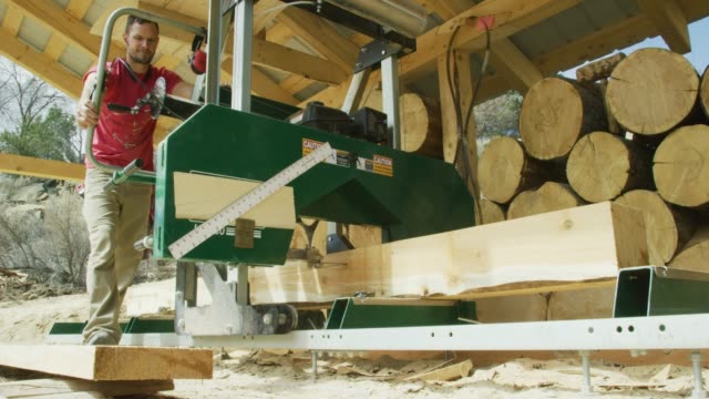 A Caucasian Man in His Thirties Uses a Log Milling Machine to Cut a Log into Wooden Planks in an Outdoor Wood Shop