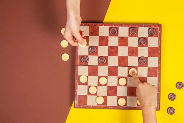 Photo of Child hands playing checkers on checker board game over yellow and blue background, top view