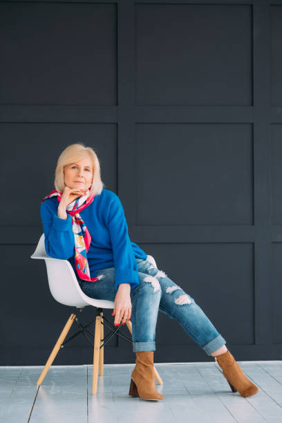 aged woman serious expression doubt trendy outfit Aged lady model. Serious facial expression. Woman sitting on modern white chair. Doubt. Casual trendy outfit. Copy space. womens blue suede shoes stock pictures, royalty-free photos & images
