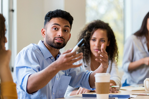 A young male design professional sits at a conference table with coworkers and gestures as he shares his ideas.