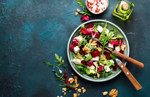 istock Beet or beetroot salad with fresh arugula, radicchio, soft cheese and walnuts on plate with fork, dressing and spices on blue kitchen table, copy space, top view 1142350519