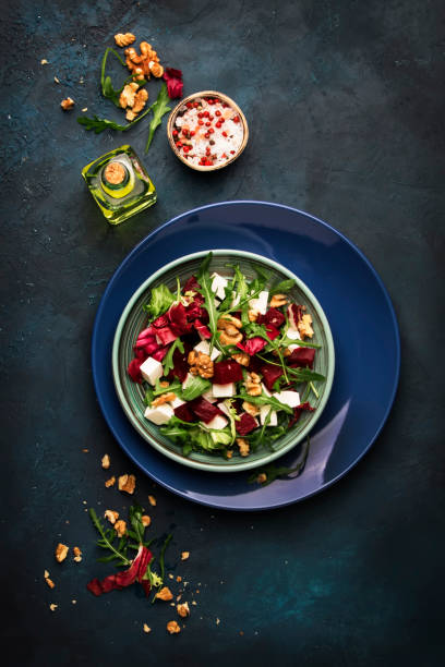 istock Beet or beetroot salad with fresh arugula, radicchio, soft cheese and walnuts on plate with fork, dressing and spices on blue kitchen table, copy space, top view 1142351030
