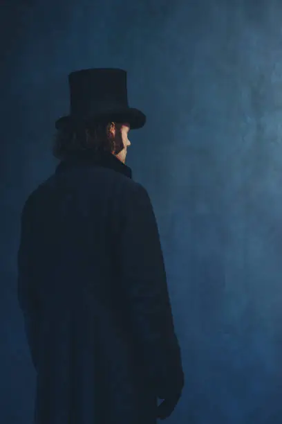 Photo of Edwardian man in long black coat and hat standing towards grey wall.