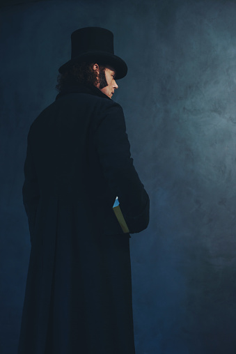 Edwardian man in long black coat and hat holding book standing towards grey wall.