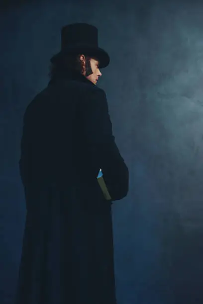 Photo of Edwardian man in long black coat and hat holding book standing towards grey wall.