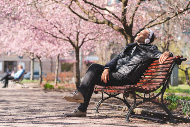 Mature man listening to music in the park A mature man is sitting on a park bench in a public park. He is enjoying the sun and listening to music, surrounded by blooming cherry blossom. one mature man only audio stock pictures, royalty-free photos & images