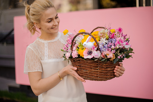 Smiling girl holding a basket of yellow and pink flowers decorated with little toy bunny especially for the Easter holiday