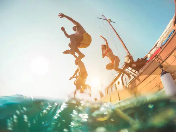 Photo of Happy friends diving from sailing boat into the sea - Young people jumping inside ocean in summer excursion day - Vacation, youth and fun concept - Main focus on close-up man - Fisheye lens distortion