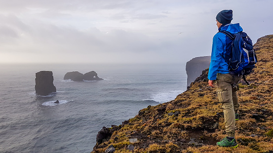 A young man wearing blue jacket, standing at the edge of a steep cliff. Some rock formations poking out of the sea. Moody atmosphere. Pure happiness and enjoyment derived from traveling