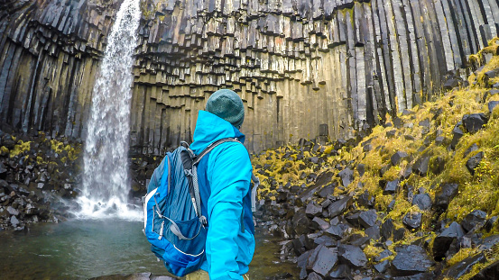 A young man wearing hiking backpack, standing in front of a waterfall, takes a selfie. Rocks on the ground look slippery. The water falls from a rock formation, decorated with moss. Svartifoss