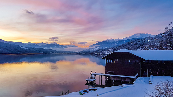 A magical sunset over the lake in Alps. Sky is pink, orange, yellow and blue. On the side there is a small hut, all covered by snow. Winter wonderland with tall snowy mountains in the back.