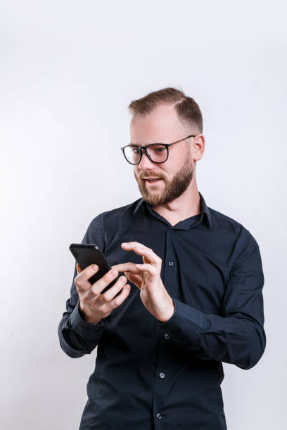 istock Young bearded man in glasses, holds modern smart phone, happy to recieve message, dressed in casual black shirt, 1142418607