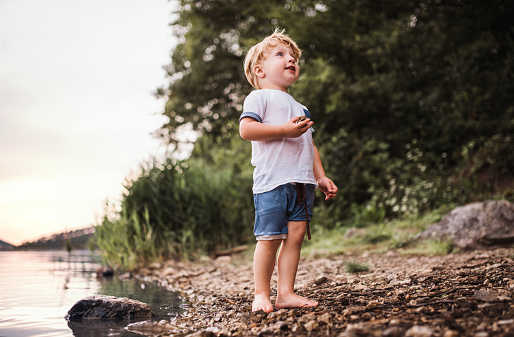 A wet, small toddler boy standing barefoot outdoors by a river in summer, playing with rocks.