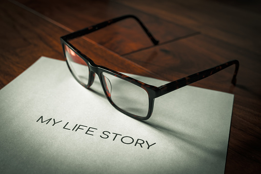 My life story wording printed on a sheet of paper sitting on a walnut floor highlighted with sunlight and a pair of spectacles in background in shallow depth of field