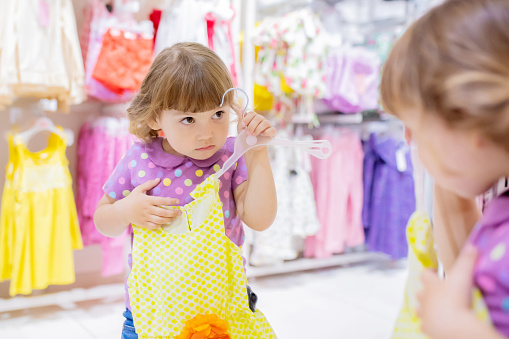 Adorable smiling little girl at the clothes store, try on new yellow summer dress, looking at the mirror.