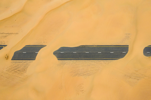 View from above, stunning aerial view of a camel crossing a deserted road covered by sand dunes. Empty road that run through the Dubai desert during sunset. Dubai, United Arab Emirates.