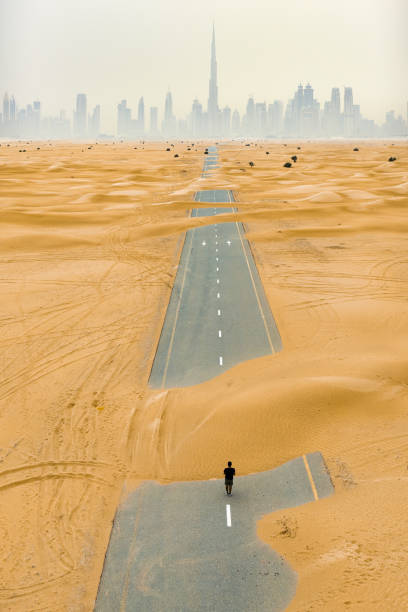 splendida vista aerea di una persona non identificata che cammina su una strada deserta coperta da dune di sabbia nel deserto di dubai. skyline di dubai circondato dalla nebbia sullo sfondo. dubai, emirati arabi uniti. - fog desert arabia sunset foto e immagini stock