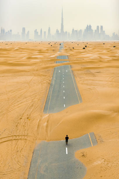 stunning aerial view of an unidentified person walking on a deserted road covered by sand dunes in dubai desert. dubai skyline surrounded by fog in the background. dubai, united arab emirates. - fog desert arabia sunset imagens e fotografias de stock