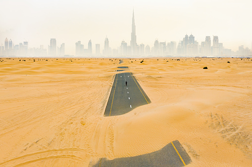 Stunning aerial view of an unidentified person walking on a deserted road covered by sand dunes in Dubai desert. Dubai skyline surrounded by fog in the background. Dubai, United Arab Emirates.