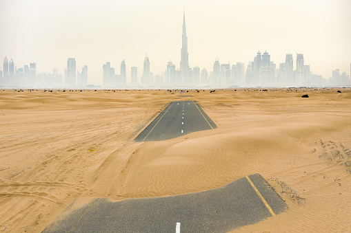 View from above, stunning aerial view of a deserted road covered by sand dunes in the middle of the Dubai desert. Beautiful Dubai skyline surrounded by fog in the background. Dubai, United Arab Emirates.