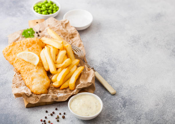 istock Traditional British Fish and Chips with tartar sauce on chopping board with fork and green peas on white stone table background. Space for text 1142440049