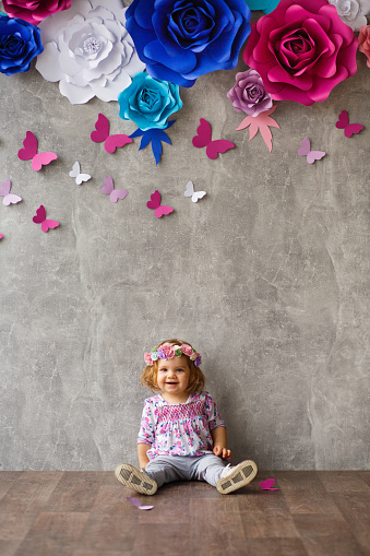 Portrait of cute baby girl with flower crown sitting against gray wall with paper flowers arrangement on it. She is looking at camera.
